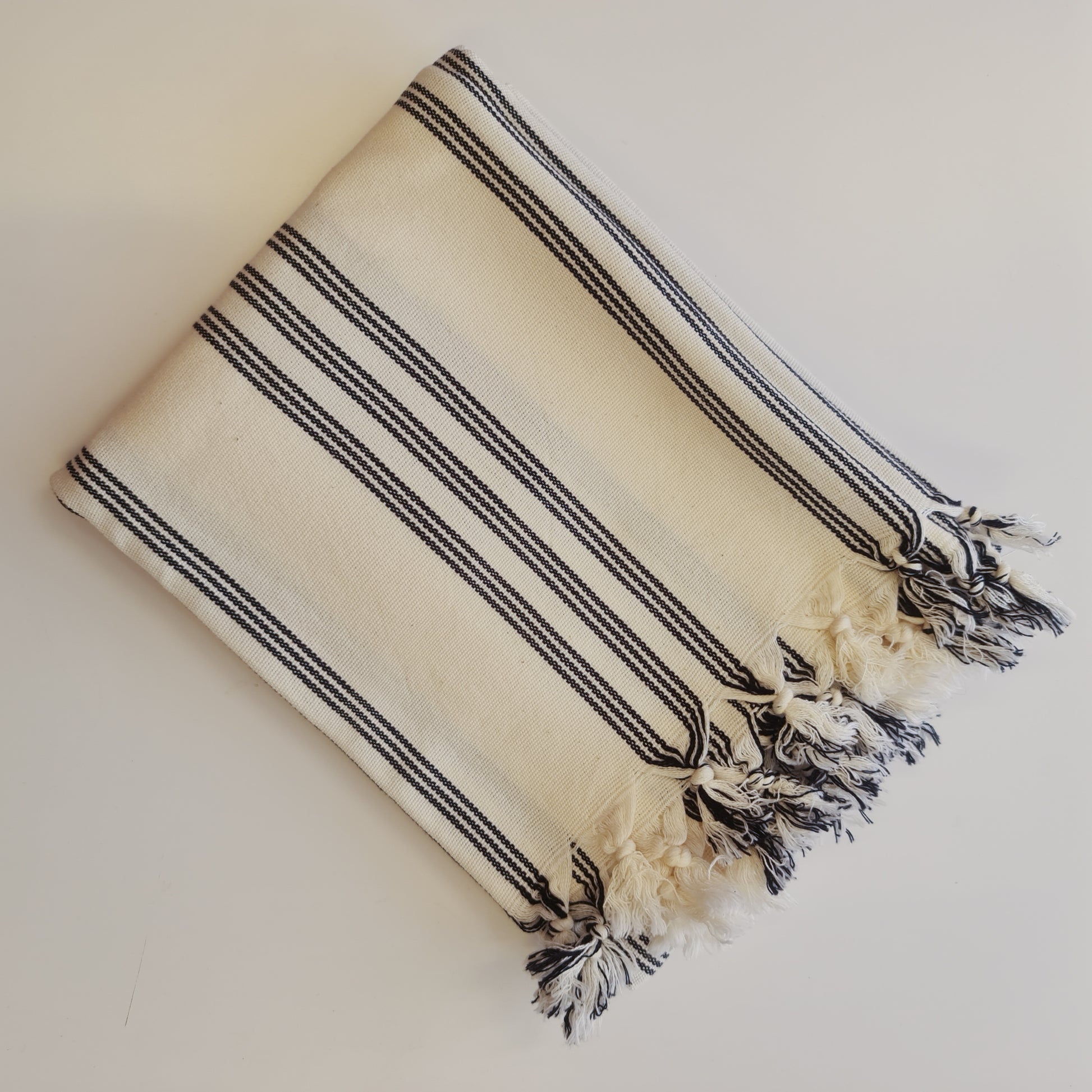 A Turkish towel with ecru tones and black stripes on a white background, with fringes on the ends.