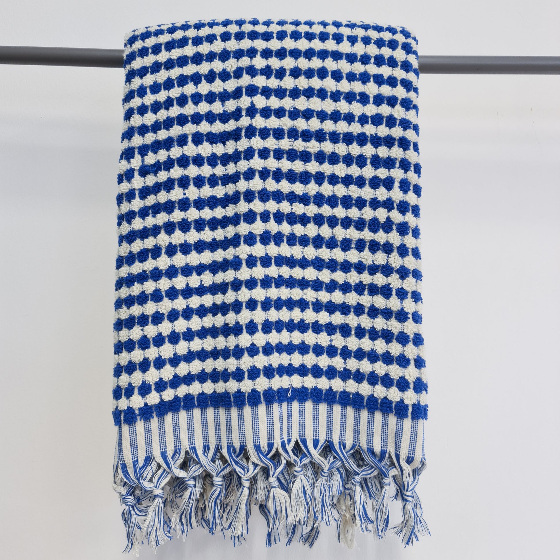 A Turkish indigo-colored terry towel with white dotted pattern, displayed on a grey rail, featuring fringes on the ends.