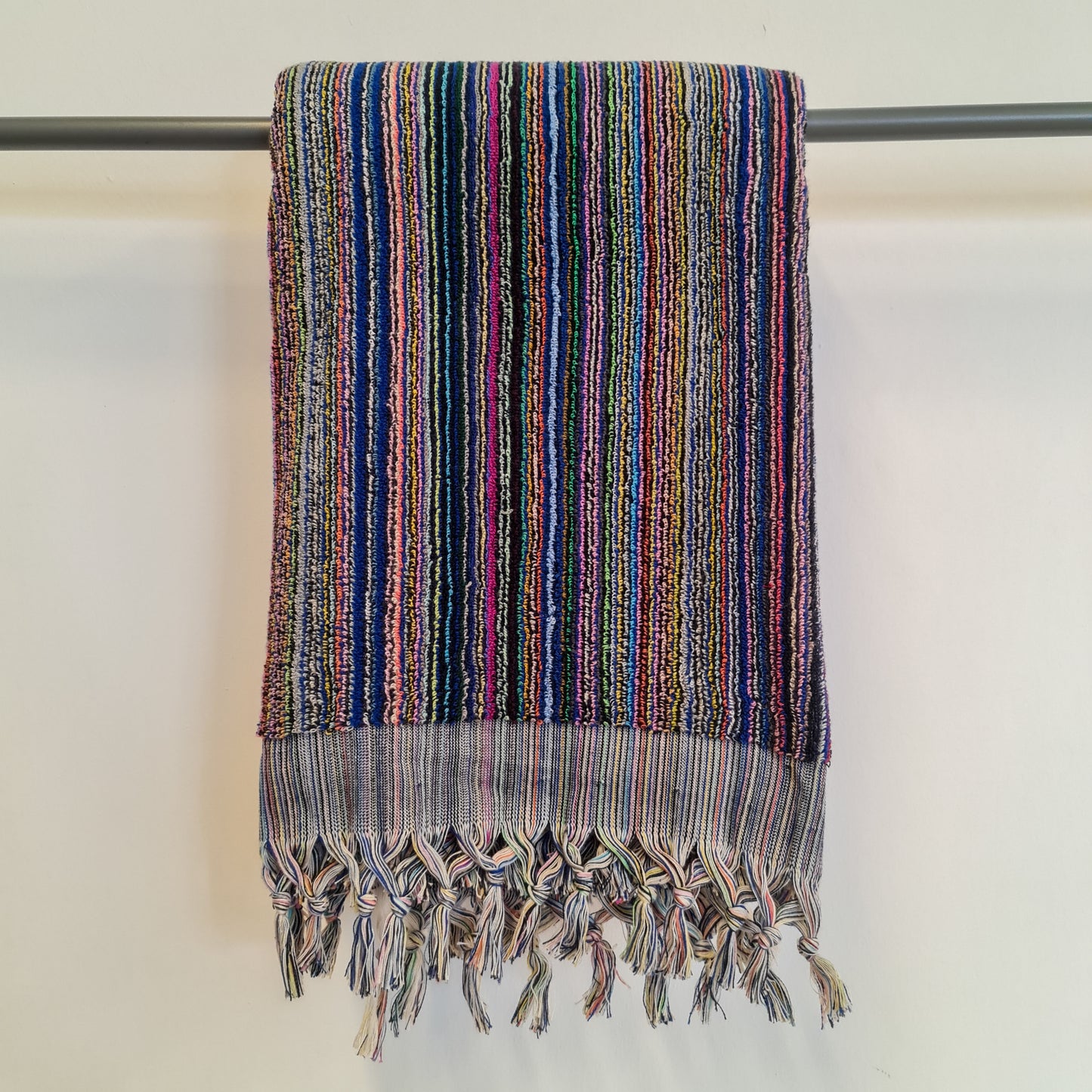 A Turkish hammam bath towel with multi-colored indigo stripes, displayed on a grey rail, featuring terry weaving on both sides.